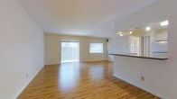 This single-floor residence offers two bedrooms and two bathrooms within a generously proportioned open-concept layout. The living area is a standout feature, highlighted by smooth hardwood-style flooring that runs throughout, crisp white walls, and large sliding glass doors paired with a full-width window that flood the space with natural light. Vertical blinds provide privacy and light control as needed. The open floor plan allows the living area to flow seamlessly toward the kitchen, which is separated by a raised breakfast bar with a dark stone-style countertop. Track lighting above the kitchen area ensures practical illumination. The neutral color palette of warm white tones and light wood flooring creates a bright, airy ambiance throughout. The layout is well-suited for comfortable everyday living, with the single-floor design offering convenient accessibility. The overall feel of the space is clean, open, and inviting, making it a functional and versatile home.
