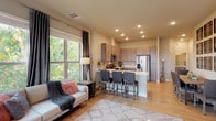 This single-floor residence offers a thoughtfully designed open-concept layout that seamlessly connects the living area and kitchen. The living room is bathed in natural light through large windows dressed with elegant floor-to-ceiling curtains, offering pleasant outdoor tree views. Light hardwood flooring flows throughout the main living areas, adding warmth to the neutral color palette.

The kitchen is outfitted with taupe shaker-style cabinetry, stainless steel appliances including a refrigerator with water dispenser, electric range, and over-the-range microwave, all complemented by clean white countertops. A peninsula with bar seating provides a casual dining option and enhances the open feel of the space.

The primary bedroom features plush carpeting, a ceiling fan, and a window with outdoor views, while a second bedroom is equally comfortable with generous natural light. Both bathrooms are well-equipped — one features a tub/shower combo and access to a walk-in closet, while the other includes an extended vanity with ample cabinetry and a linen tower for additional storage. Recessed lighting throughout the home ensures a bright and welcoming ambiance across every room.
