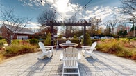 Located at 10035 Dabney Dr in Charlotte, NC, this well-appointed outdoor amenity space offers a thoughtfully designed single-level patio environment ideal for relaxation and social gatherings. The space is anchored by a stylish pergola structure with dark wood beams and strung café lights, creating an inviting ambiance during both daytime and evening hours.

A standout feature is the stacked-stone bar counter topped with a sleek concrete surface, accompanied by high-back bar seating — perfect for casual dining or entertaining. Adjacent to the bar area, a separate pergola-shaded lounge zone centers around a circular fire pit table, surrounded by classic white Adirondack chairs arranged to encourage conversation and outdoor leisure.

The expansive concrete patio connects multiple zones seamlessly, while additional outdoor seating areas with standard patio furniture provide further flexibility for gathering. The surrounding landscaping features a curated mix of ornamental grasses, evergreen shrubs, columnar trees, and seasonal plantings that add color and texture throughout the year.

A nearby brick building with traditional shuttered windows and well-maintained garden beds completes the setting, contributing to the overall charm and character of the space. The combination of functional amenities, durable materials, and attractive greenery makes this outdoor space a welcoming retreat in the Charlotte community.