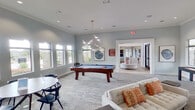 This contemporary single-floor clubhouse amenity space is thoughtfully designed to offer a well-rounded social and recreational experience. The main lounge area is an expansive, open space finished in soft neutral tones, anchored by a full-size pool table and a sculptural geometric chandelier that serves as a striking focal point. Plush tufted sofas with warm accent pillows, a shuffleboard table, and a wall-mounted flat-screen TV complete the entertainment offerings, while large windows along multiple walls invite generous natural light and views of the surrounding greenery. Curated abstract artwork is displayed throughout, adding a refined artistic touch to the atmosphere. Adjacent to the lounge, a spacious kitchen bar area features a long white quartz countertop island, warm wood-look flooring, globe pendant lighting, and a wall of windows that floods the space with light, making it ideal for casual gatherings. A dedicated wet bar area is highlighted by a bold geometric-patterned tile accent wall with sleek cabinetry and dual faucet stations. A private conference room offers a glass-top table with modern leather chairs and natural light from street-facing windows. The space flows seamlessly between zones, balancing function with a polished, modern aesthetic throughout.