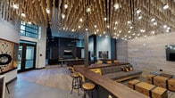 Located at 430 Jackson St in Oakland, CA, this single-floor community lounge is a masterclass in industrial-modern design. The expansive main area commands attention with soaring double-height ceilings draped in a dramatic installation of hanging rope and Edison bulb pendants, casting a warm, ambient glow throughout the space. Wide-plank hardwood flooring runs seamlessly underfoot, complemented by light wood-paneled accent walls that add warmth and texture.

The lounge is thoughtfully divided into distinct social zones. A game area features a full-size shuffleboard table and a billiards table, while a lounge seating zone offers plush sofas and armchairs accented with bold yellow throw pillows, centered around a wall-mounted television and sleek fireplace. High-top communal tables with industrial-style stools provide additional gathering space, ideal for casual conversation or co-working.

The fully equipped kitchen area is a standout feature, showcasing crisp white countertops, matte black cabinetry, a metallic tile backsplash, a professional-grade gas range, and a draft beer tap — all framed by a chalkboard beer-themed mural. A large central island anchors the space.

Large glass double doors open to an outdoor courtyard, flooding the interior with natural light. A perforated black sliding barn door adds an artful industrial touch at the entrance corridor, where Oakland-themed photography murals pay homage to the city's vibrant character.