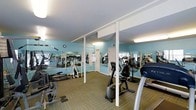 This fitness center offers a spacious, single-floor layout designed to accommodate a variety of workout routines. The room is finished with light blue walls that create a fresh, energizing atmosphere, complemented by a white ceiling with recessed and fluorescent lighting that ensures the space is consistently well-lit throughout. Large mirrors line the walls, visually expanding the room and allowing users to monitor their form during exercise. The flooring consists of patterned carpet tiles in earthy tones, providing both comfort underfoot and a practical surface for the training environment. The facility is equipped with a range of cardio machines, including stationary bikes, treadmills, and elliptical trainers, as well as strength and resistance equipment positioned throughout the floor. White structural columns are integrated into the layout, adding architectural definition without disrupting the flow of the space. A wall clock is mounted for easy time-keeping during workouts. The overall design prioritizes functionality and openness, making it a practical and accessible fitness destination.