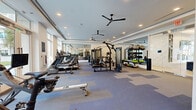 Located at 111 Green St SE in Gainesville, Georgia, this single-floor fitness center is designed to deliver a comprehensive, upscale workout experience. The facility's layout is expansive and open, with tray ceilings, recessed lighting, and multiple ceiling fans contributing to a clean, modern aesthetic throughout.

The main exercise floor is generously proportioned, accommodating a wide array of cardio equipment along one side, including treadmills, ellipticals, rowing machines, and stationary bikes. Floor-to-ceiling windows line the perimeter, flooding the space with abundant natural light and providing views of the surrounding outdoor courtyard and streetscape. Full-length mirrors are strategically positioned to enhance the sense of depth and openness.

The strength training area features multi-functional cable machines, a functional trainer, vertical knee raise stations, and a well-stocked rack of stability balls and kettlebells. A dedicated free-weight section with benches and a dumbbell rack rounds out the strength offerings. A water fountain station and cubbies provide added convenience for users.

The facility is accessed through a sophisticated lobby entry featuring light hardwood flooring, a dramatic marble-clad accent wall, glass partitions, and elegant pendant lighting, setting a refined tone from the moment of arrival.