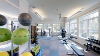 Located at 1910 Capital Creek Dr in Wake Forest, NC, this fitness center offers a bright, well-appointed single-floor exercise space designed to support a wide range of workout routines. Large windows line the walls, flooding the room with abundant natural light and providing views of the surrounding outdoor scenery, creating an energizing atmosphere for exercise. The space is finished with a durable, multi-toned blue and gray carpet tile flooring that adds both comfort and visual appeal underfoot. The ceiling is outfitted with a ceiling fan and recessed lighting fixtures, ensuring proper air circulation and illumination throughout the day. The facility is comprehensively equipped with strength training machines, a functional trainer cable system, and adjustable benches to accommodate various resistance exercises. A dedicated functional training area features an organized rack stocked with resistance bands, suspension trainers, stability balls in multiple sizes, medicine balls, and rolled exercise mats, supporting dynamic and group fitness activities. The clean, white-painted walls and neutral trim give the space a modern, professional aesthetic. The open layout allows for ample movement between equipment stations, making the facility accessible and practical for users of all fitness levels.