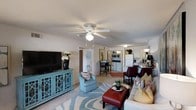 This single-floor residence features an open-concept floor plan that seamlessly connects the living, dining, and kitchen areas, creating a cohesive and functional living environment. The living area is generously sized, anchored by a bold turquoise media console housing a large flat-screen television, and complemented by plush seating including a light blue accent chair and a neutral sofa adorned with colorful throw pillows. A stylish patterned area rug in blue and white tones grounds the space, while a ceiling fan with integrated lighting provides both comfort and illumination.

The open sightlines lead directly into the dining area, which accommodates a small table with bar-height seating, and further into the kitchen, which features white cabinetry, a refrigerator, microwave, and ample counter space. The neutral wall tones and light-colored flooring throughout contribute to a bright and airy atmosphere. The layout maximizes the use of space, making it both practical and visually inviting, with a consistent design aesthetic carried through each connected area.