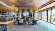 This sophisticated single-floor amenity space offers a generously sized, open-concept layout thoughtfully designed for both social gathering and relaxed lounging. The interior is unified by a striking combination of textured wood-slat ceilings, warm amber woven pendant lights, and mixed flooring that transitions between large-format tile and light hardwood, creating distinct yet cohesive zones throughout.

The living lounge area serves as the centerpiece, featuring plush seating arrangements, a bold decorative lattice screen partition, and large-scale artwork. Floor-to-ceiling glass sliding doors flood the space with natural light and provide seamless access to the outdoor patio, where additional seating and open-air views await.

Adjacent to the lounge is a dedicated billiards area, complete with a full-sized pool table and stylish accent furnishings, perfect for entertainment. A fully equipped kitchen and bar zone anchors the space with a long granite-topped island, leather-upholstered bar stools, warm gold ceilings, stone tile backsplash, built-in appliances, and dual wall-mounted flat-screen televisions — ideal for hosting and casual dining.

A dedicated dining area with round tables, patterned upholstered chairs, and additional pendant lighting rounds out the interior. Together, these spaces create a vibrant, well-appointed environment that balances comfort, function, and contemporary design.