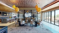 This sophisticated single-floor amenity space offers a generously sized, open-concept layout thoughtfully designed for both social gathering and relaxed lounging. The interior is unified by a striking combination of textured wood-slat ceilings, warm amber woven pendant lights, and mixed flooring that transitions between large-format tile and light hardwood, creating distinct yet cohesive zones throughout.

The living lounge area serves as the centerpiece, featuring plush seating arrangements, a bold decorative lattice screen partition, and large-scale artwork. Floor-to-ceiling glass sliding doors flood the space with natural light and provide seamless access to the outdoor patio, where additional seating and open-air views await.

Adjacent to the lounge is a dedicated billiards area, complete with a full-sized pool table and stylish accent furnishings, perfect for entertainment. A fully equipped kitchen and bar zone anchors the space with a long granite-topped island, leather-upholstered bar stools, warm gold ceilings, stone tile backsplash, built-in appliances, and dual wall-mounted flat-screen televisions — ideal for hosting and casual dining.

A dedicated dining area with round tables, patterned upholstered chairs, and additional pendant lighting rounds out the interior. Together, these spaces create a vibrant, well-appointed environment that balances comfort, function, and contemporary design.