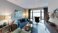 This contemporary single-floor residence showcases a thoughtfully designed layout that balances modern aesthetics with everyday functionality. The kitchen is a standout feature, outfitted with high-gloss white upper cabinetry, dark lower cabinets, stainless steel appliances including a French door refrigerator and range with hood, white quartz countertops, and a textured stone tile backsplash. A generous island provides additional prep and serving space, all illuminated by track lighting and pendant fixtures that enhance the clean, modern look. Light wood-toned flooring flows throughout, lending warmth to the crisp design palette.

The living area is bright and inviting, anchored by floor-to-ceiling windows and a sliding glass door that open to a private balcony, allowing natural light to pour in and offering views of the surrounding streetscape. The space accommodates comfortable seating arrangements and features bold, colorful artwork that adds personality to the neutral gray walls.

The bedroom continues the contemporary theme, highlighted by a dramatic four-poster canopy bed frame, warm golden drapery framing large windows, and direct access to the balcony. A rustic wood dresser with a round mirror adds a contrasting organic touch.

The full bathroom features large-format subway tile surrounding the soaking tub and shower combo, accented by a dark tile border, gray floor tile, and a modern vanity, completing this well-curated residence.