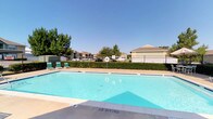 Located in Sherman, this community pool area offers a well-maintained outdoor retreat designed for relaxation and recreation. The centerpiece is a generously sized swimming pool with clear, blue water, marked with depth indicators and equipped with safety features including a life ring station. The pool deck is finished in a light-colored concrete surface, providing ample space for lounging and movement around the pool.

Surrounding the pool, multiple reclining lounge chairs and chaise lounges are positioned to take advantage of the open sky and abundant natural sunlight. A covered patio area with a shade umbrella and additional seating provides a comfortable spot to relax out of direct sun. The entire area is enclosed by a sturdy metal perimeter fence, ensuring a secure environment.

Adjacent to the pool is a single-story clubhouse building featuring a pitched shingle roof, covered entryway, and large windows. The building includes amenity stations such as a vending machine visible from the pool deck. Mature trees are scattered throughout and beyond the fenced area, offering natural shade and greenery. An American flag flies prominently near the clubhouse, adding to the community character of this inviting outdoor space.