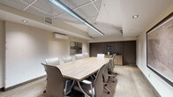 Located at 1100 Avenue du Docteur-Penfield in Montréal, this professional conference room offers a polished and functional environment designed for productive meetings. The room centers around a spacious light wood boardroom table surrounded by ergonomic high-back chairs upholstered in neutral tones, comfortably accommodating multiple attendees. The ceiling features a combination of a decorative perforated tile system and recessed lighting, complemented by a prominent linear LED fixture that runs the length of the table, ensuring well-distributed illumination throughout the space. A wall-mounted mini-split air conditioning unit provides climate control, contributing to a comfortable working environment year-round. The walls are finished in a warm neutral palette, with one side adorned by an abstract canvas artwork and another featuring a framed vintage map of the City of Montréal, adding character and a sense of place. A built-in wood-toned credenza along one wall provides practical storage, while a wall-mounted whiteboard offers a tool for collaboration and presentations. Textured carpet tiles in a subtle pattern cover the floor, and a solid wood door with warm tones provides a refined entry point. The room is equipped with a display screen and security camera, making it a fully functional, professional meeting space.