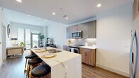 This thoughtfully designed two-bedroom, one-bathroom residence in Silver Spring, Maryland offers a modern open-concept layout filled with contemporary finishes and natural light. The kitchen is a standout feature, boasting a striking waterfall-edge quartz island with bar seating, two-tone cabinetry in warm wood and matte gray, a subway tile backsplash, stainless steel appliances, and hardwood-style flooring that flows seamlessly into the living area. The bright living room opens directly onto a private balcony through a full-height glass door, offering sweeping outdoor views and abundant natural light. The primary bedroom is generously proportioned, featuring plush carpeting, large windows with outdoor views, and a clean, airy aesthetic. The second bedroom is equally well-appointed. A hallway connects the bedrooms to the shared full bathroom, which features marble-style tile flooring, a wide vanity with an undermount sink, and a shower-tub combination. A dedicated closet provides additional storage. Recessed lighting throughout, crisp white walls, and a cohesive modern palette give the entire residence a polished, contemporary feel.