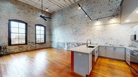 This two-floor space showcases a skillful adaptive reuse of an industrial building, preserving its historic character while incorporating contemporary upgrades throughout. The main level is anchored by a generously scaled open-plan living and kitchen area defined by soaring ceilings with whitewashed exposed brick walls, original painted-wood beam ceilings, and warm reclaimed hardwood flooring. Track lighting and a ceiling fan complement the arched multi-pane windows that draw in natural light. The kitchen features a bold red-accented island topped with granite countertops, gray shaker cabinetry with black hardware, a tile backsplash, and stainless steel appliances. A corridor on this level leads to additional rooms and a bathroom equipped with a pedestal sink and dark-framed fixtures. The upper level continues the reclaimed hardwood flooring and offers a loft-style layout with open railings overlooking the lower floor. An exposed structural ceiling with painted beams maintains the industrial aesthetic. The upper floor also includes a bedroom with an en-suite bathroom featuring gray vanity cabinetry, as well as a walk-in closet. Throughout both levels, the interplay of raw brick, bold structural columns, and refined modern finishes creates a distinctive and cohesive industrial-chic atmosphere.