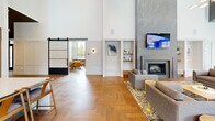 This generously sized single-floor amenity space in Indianapolis, IN (46240) offers a well-appointed, contemporary layout designed for both social gathering and relaxed lounging. Soaring ceilings—dramatically double-height in key areas—create an open, airy atmosphere throughout, while herringbone-patterned hardwood flooring provides a refined, cohesive aesthetic across all zones.

The main lounge area centers around a floor-to-ceiling stone fireplace surround with a wall-mounted television above, complemented by built-in shelving niches and wainscoting detail. Seating areas are defined by curated furniture groupings and stylish area rugs. A black-framed sliding barn door connects distinct zones while maintaining visual continuity.

The full kitchen features warm wood cabinetry, stainless steel appliances, open floating shelves, and an expansive white quartz island with seating for multiple guests—ideal for community events or casual gatherings.

A dedicated game room includes an octagonal game table, shuffleboard, and a wall-mounted TV, with floor-to-ceiling windows framing outdoor views. Additional seating nooks positioned along the perimeter offer quieter, light-filled spaces for conversation, each benefiting from large-format windows overlooking the surrounding community and pool area.