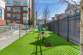 Located in Atlanta, GA, this outdoor dog park is a well-appointed amenity designed to provide a safe and engaging environment for pets and their owners. The space is enclosed by a combination of sturdy black chain-link and ornamental iron fencing, ensuring secure containment throughout. Entry is managed through a gated access point equipped with a locking mechanism, adding an additional layer of safety.

The ground is covered in lush artificial turf, providing a clean, low-maintenance surface that remains green year-round. A concrete pathway runs through portions of the space, offering a firm walking surface between areas. Agility equipment is a highlight of the park, including a tunnel and a ramp structure, both finished in green and designed to encourage active play and exercise for dogs of varying sizes.

Mature trees are planted within the enclosure, offering natural shade during sunny Atlanta days. Seating benches are positioned near the perimeter, providing comfortable resting spots for pet owners to supervise their animals. The surrounding multi-story brick residential buildings frame the space, contributing an urban backdrop while the open sky above ensures the area remains bright and well-lit throughout the day.