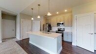 Located at 510 Morning Oak Drive in Westfield, Indiana, this single-floor residence offers a thoughtfully designed layout with contemporary finishes throughout. The kitchen serves as a standout feature, centered around a large white-topped island with a built-in sink and pendant lighting overhead. Gray shaker-style cabinetry lines the walls, complemented by a classic white subway tile backsplash, light quartz countertops, and a full suite of stainless steel appliances including a refrigerator, electric range, and over-the-range microwave. Rich dark hardwood-style flooring runs through the kitchen and adjoining areas, creating a warm contrast against the neutral wall tones. The living area transitions seamlessly from hard flooring to plush carpeting, with vaulted ceiling angles adding a sense of openness and a sliding glass door inviting natural light into the space. The bedroom continues the carpeted flooring with vaulted ceiling details and a window providing natural light and exterior views. The bathroom is well-appointed with gray cabinetry, a white vanity countertop, a tiled soaking tub and shower combination, and matching dark hardwood-style flooring, along with convenient access to what appears to be a closet or additional storage area.