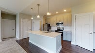 Located at 510 Morning Oak Drive in Westfield, Indiana, this single-floor residence offers a thoughtfully designed layout with contemporary finishes throughout. The kitchen serves as a standout feature, centered around a large white-topped island with a built-in sink and pendant lighting overhead. Gray shaker-style cabinetry lines the walls, complemented by a classic white subway tile backsplash, light quartz countertops, and a full suite of stainless steel appliances including a refrigerator, electric range, and over-the-range microwave. Rich dark hardwood-style flooring runs through the kitchen and adjoining areas, creating a warm contrast against the neutral wall tones. The living area transitions seamlessly from hard flooring to plush carpeting, with vaulted ceiling angles adding a sense of openness and a sliding glass door inviting natural light into the space. The bedroom continues the carpeted flooring with vaulted ceiling details and a window providing natural light and exterior views. The bathroom is well-appointed with gray cabinetry, a white vanity countertop, a tiled soaking tub and shower combination, and matching dark hardwood-style flooring, along with convenient access to what appears to be a closet or additional storage area.