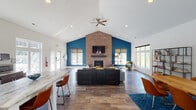 This single-floor community clubhouse is thoughtfully designed to offer a range of amenities in a welcoming, contemporary setting. The entrance foyer greets visitors with soaring vaulted ceilings, rich hardwood-style flooring, and elegant décor, setting a polished tone throughout.

The expansive main lounge is a standout feature, anchored by a full-height brick fireplace flanked by bold teal accent walls and a wall-mounted television. Vaulted ceilings with a ceiling fan amplify the sense of openness, while generous windows flood the space with natural light and outdoor views. Comfortable seating arrangements make this an ideal gathering area.

Adjacent to the lounge is a dining area furnished with a large wooden table and leather chairs, accented by decorative wall art and a vibrant blue area rug. A fully equipped kitchen with dark cabinetry, stainless steel appliances, and marble-style countertops supports community events and gatherings.

The dedicated fitness center, branded "Get Fit," is well-appointed with cardio machines including treadmills, ellipticals, and a stationary bike, as well as strength equipment, free weights, and wall-mounted televisions. Large windows provide natural light and views of the outdoor pool area, creating an energizing workout environment. Hardwood-style flooring and full-length mirrors complete the space.