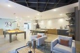 Located at 3050 Circle 75 Pkwy SE in Atlanta, Georgia, this single-floor amenity lounge is a well-appointed communal space designed for socializing and recreation. The layout encompasses several distinct zones, each thoughtfully curated with contemporary finishes and furnishings.

The lounge area serves as a central gathering space, featuring a sleek wall-mounted flat-screen television above a linear fireplace set against dark, textured wall panels. A coffered black ceiling adds dramatic depth overhead, while large windows along one side offer views of an outdoor pool area, filling the space with natural light. Comfortable cream-toned seating arranged around a wood-and-metal coffee table completes the lounge setting.

Adjacent to the lounge, a wet bar and service counter stretches along one wall, highlighted by a striking floor-to-ceiling marble slab backsplash with bold veining. The counter features warm wood cabinetry, a double sink, beverage tap system, and under-counter refrigeration, making it well-suited for entertaining.

A billiards table anchors the game area, providing a casual recreational focal point. The dining nook zone offers booth-style seating with solid wood dividers, white tabletops, upholstered benches, and sculptural pendant lighting against soft blue-gray walls, completing this versatile and stylish communal retreat.