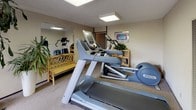 This fitness center is a single-level exercise space designed to accommodate essential cardio and workout needs. The room features wall-to-wall carpeting and a textured ceiling with recessed flush-mount lighting panels that provide even, functional illumination throughout the space. A large mirror spans one wall, visually expanding the room and allowing users to monitor their form during workouts.

The equipment lineup includes treadmills and elliptical trainers from Precor, a recognized fitness equipment brand, positioned to make efficient use of the available floor space. Wooden shelving units along the walls hold potted plants, adding a touch of greenery and a welcoming atmosphere to the otherwise utilitarian setting. A rattan bench is also present, offering a place to rest or store personal belongings, and magazines are stored nearby for convenience.

Large sliding glass doors on one wall open directly to an outdoor patio area furnished with wrought-iron table and chairs beneath a striped umbrella, seamlessly connecting the interior workout space with an exterior relaxation area. The natural light that filters through these doors brightens the room and provides views of the surrounding landscaped courtyard, enhancing the overall experience of the fitness center.
