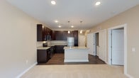This well-appointed single-floor residence showcases a thoughtfully designed open-concept layout that seamlessly blends the kitchen and living areas. The kitchen is a standout feature, boasting rich dark espresso cabinetry that extends to the ceiling, offering generous storage throughout. Light granite countertops provide a striking contrast against the dark cabinetry, while a central island with matching countertops serves as both a functional prep space and a casual gathering spot. Stainless steel appliances, including a refrigerator, range, and over-the-range microwave, lend a sleek, contemporary feel to the space. Three elegant pendant lights hang above the island, complementing the recessed lighting overhead to create a warm, well-lit environment. The kitchen transitions smoothly into the living area, where plush carpeting provides a comfortable and inviting atmosphere. Dark hardwood-style flooring in the kitchen ties the design together with a cohesive aesthetic. The neutral wall tones throughout enhance the sense of openness, while multiple doorways visible from the main space suggest convenient access to additional rooms, making this a highly functional and visually appealing home.
