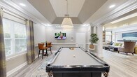 Located at 2600 Midway Branch Dr in Odenton, Maryland, this well-appointed community amenity space offers a variety of functional and social areas across a single, generously sized floor. The entrance foyer sets a refined tone with large-format tile flooring, a bold sputnik-style chandelier, framed artwork, and a striking round pedestal table as a centerpiece.

The game room is a standout feature, highlighted by a vaulted tray ceiling, a pendant light fixture, and a full-size pool table positioned atop a patterned area rug. A wall-mounted flat-screen TV and a bistro-height bar table with leather chairs complete the space, which opens seamlessly into the adjacent lounge area through decorative columned archways.

The lounge exudes comfort and style, featuring rich crown molding, a large sectional sofa adorned with colorful accent pillows, curated artwork, and lush indoor greenery. Light wood-look flooring flows throughout, unifying the spaces. Wainscoting with beadboard paneling adds a classic architectural touch to the corridors and transitional areas.

A compact kitchenette offers granite countertops, warm wood cabinetry, stainless steel appliances, and decorative wall art. The business center provides a practical workstation with granite desk surfaces, wood-look flooring, and access to an exterior glass door, making it a versatile and well-equipped workspace.