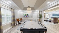 Located at 2600 Midway Branch Dr in Odenton, Maryland, this well-appointed community amenity space offers a variety of functional and social areas across a single, generously sized floor. The entrance foyer sets a refined tone with large-format tile flooring, a bold sputnik-style chandelier, framed artwork, and a striking round pedestal table as a centerpiece.

The game room is a standout feature, highlighted by a vaulted tray ceiling, a pendant light fixture, and a full-size pool table positioned atop a patterned area rug. A wall-mounted flat-screen TV and a bistro-height bar table with leather chairs complete the space, which opens seamlessly into the adjacent lounge area through decorative columned archways.

The lounge exudes comfort and style, featuring rich crown molding, a large sectional sofa adorned with colorful accent pillows, curated artwork, and lush indoor greenery. Light wood-look flooring flows throughout, unifying the spaces. Wainscoting with beadboard paneling adds a classic architectural touch to the corridors and transitional areas.

A compact kitchenette offers granite countertops, warm wood cabinetry, stainless steel appliances, and decorative wall art. The business center provides a practical workstation with granite desk surfaces, wood-look flooring, and access to an exterior glass door, making it a versatile and well-equipped workspace.
