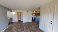 This single-story unit is thoughtfully laid out across a compact, efficient floor plan. The open living area features dark hardwood-style flooring and a textured ceiling, with a ceiling fan providing additional airflow. Natural light filters in through vertical-blind-covered windows and a sliding glass door, lending the space a bright, airy feel.

The kitchen is positioned adjacent to the living area and is defined by a teal-accented peninsula with a light-toned countertop, offering both additional workspace and a casual dining or serving surface. Gray cabinetry lines the walls above and below the counters, providing generous storage. White appliances, including a refrigerator, electric range, and an under-cabinet range hood, complete the functional cooking space. A double-basin stainless steel sink is integrated into the countertop.

The bedrooms are carpeted in neutral tones, each featuring windows with vertical blinds that allow natural light into the rooms. One bedroom includes a closet accessible through a dedicated doorway with a walk-in configuration. The bathroom is fitted with a full tub and shower combo accented with white tile, chrome fixtures, and a towel bar. Textured ceilings are consistent throughout the unit, contributing to a cohesive interior aesthetic.