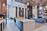 Located at 900 Battery Ave SE in Atlanta, Georgia, this single-floor clubhouse lobby presents a refined and thoughtfully designed communal space. Soaring double-height ceilings immediately establish a sense of grandeur, while large floor-to-ceiling windows and glass entry doors flood the interior with natural light and frame views of the outdoor pool area beyond.

The main lounge area anchors the space with a tufted leather sofa, layered with rich navy and earth-toned accent pillows, arranged atop a patterned area rug. Cylindrical glass pendant lights hang at varying heights from the ceiling, casting a warm, ambient glow over the seating and service bar. A dedicated coffee and beverage bar with a dark countertop and pendant lighting serves as a functional focal point.

Multiple distinct seating vignettes offer versatile settings throughout the space. A pair of deep green velvet chairs flank a sculptural side table beneath a darkly painted accent wall adorned with circular mirrors and curated photography. Bistro-style café tables with black Windsor chairs are positioned against an olive-toned accent wall showcasing large-scale abstract artwork in bold brushstrokes of gold, black, and white.

Large-format porcelain tile flooring transitions seamlessly throughout, complemented by wood accents and a neutral architectural palette that ties each zone together into a cohesive, upscale environment.