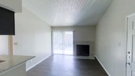 This single-floor space presents a clean, functional living area designed with a neutral, modern aesthetic. The main living space features smooth, dark hardwood-style flooring that contrasts cleanly against the light gray walls and white baseboards, creating a contemporary yet understated look. A textured popcorn ceiling adds a classic touch overhead.

A standout feature of the living area is the tiled fireplace, positioned along one wall and framed with gray tile surround, offering both a functional and visual focal point to the room. Adjacent to the fireplace, a sliding glass door fitted with vertical blinds allows an abundance of natural light to fill the space, while also providing direct access to an outdoor area, suggesting a patio or balcony beyond. The outdoor greenery visible through the glass adds a welcoming sense of connection to the exterior environment.

The open layout of the living area gives the space a spacious, airy feel, with minimal obstructions allowing for flexible furniture arrangement. Overall, the space presents a well-maintained, move-in-ready environment with practical features and a calm, neutral palette throughout.