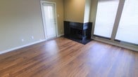 This single-floor space offers a clean, functional layout with a warm and inviting aesthetic. The living area is anchored by rich, wide-plank hardwood flooring that runs throughout, adding warmth and character to the space. A striking black-tiled fireplace serves as the room's focal point, featuring a full tile surround that extends to the floor, creating a bold contrast against the neutral, warm-toned walls. The fireplace is positioned as a corner-adjacent feature, giving it a commanding presence without overwhelming the open feel of the room. Natural light flows generously into the space through multiple windows fitted with horizontal blinds, allowing for flexible light control throughout the day. A glass-paneled door provides additional light and offers access to an exterior area. The walls are finished in a soft, earthy tone that complements the wood flooring and ties the overall palette together cohesively. White baseboards and door trim provide crisp definition throughout. The space feels open and well-proportioned, with a straightforward layout that is easy to navigate. Overall, the combination of hardwood flooring, a bold fireplace feature, and ample natural light gives this space a comfortable, classic character.