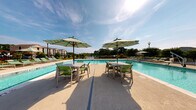 Located at 6710 Wahoo Drive in Myrtle Beach, South Carolina, this outdoor amenity space offers a well-appointed resort-style experience designed for relaxation and social gathering. At the heart of the space is a generously sized swimming pool surrounded by a broad concrete deck furnished with dining tables, chairs, and striped umbrellas, providing ample shade on sunny days. The pool area benefits from picturesque views of a neighboring pond and lush mature trees, creating a serene natural backdrop.

A covered patio runs alongside the clubhouse building, featuring a warm wood-paneled ceiling, ceiling fans, and open-air seating that includes wicker sofas, armchairs, and coffee tables arranged for comfortable lounging. A wall-mounted flat-screen television adds an entertainment element to this outdoor living area. The covered corridor connects seamlessly to the interior through double glass doors framed by classic brick accents and green lap siding, reflecting a coastal-inspired architectural style.

Wrought iron fencing and gated access define the transition between the covered patio and the pool deck, ensuring a secure and well-organized environment. The space enjoys abundant natural light throughout the day, with open views of the surrounding residential community and landscaped grounds, making it an inviting retreat for residents seeking both activity and relaxation.