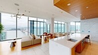 The Skylounge at 399 Fremont is a 1,780-square-foot premium amenity space located in San Francisco, CA 94105, offering breathtaking, unobstructed views of the San Francisco Bay, the iconic Bay Bridge, and the surrounding cityscape through floor-to-ceiling glass curtain walls that wrap the perimeter of the space.

The interior is defined by a refined, contemporary design palette. Large-format light stone tile flooring runs throughout, complemented by warm bamboo wood accents and crisp white surfaces. A dramatic full-height marble feature wall serves as a central architectural focal point, its veined white stone adding texture and elegance to the open layout.

The lounge area is furnished with plush, neutral-toned sectional sofas, sculptural accent chairs in warm cognac tones, and minimalist side tables, all arranged to maximize enjoyment of the sweeping views. Designer pendant chandeliers and branching chandelier fixtures provide both ambient and accent lighting, while recessed cove lighting with warm amber accents along the ceiling adds a sophisticated, atmospheric glow.

A built-in bar and counter area features a white waterfall-edge island with bar seating, warm wood cabinetry, and a mounted flat-screen display — ideal for entertaining. The space also includes a quieter lounge alcove, offering a more intimate setting. Together, these areas create a versatile, upscale environment suited for social gatherings or relaxed retreat.