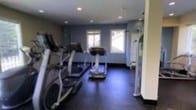 This single-floor fitness facility is designed to provide a functional and focused workout environment. The space is outfitted with multiple cardio machines, including treadmills and an elliptical trainer, positioned to make efficient use of the available floor area. The dark flooring provides a durable, gym-appropriate surface that contrasts with the cool blue-grey tones of the surrounding walls, lending the room a clean, modern aesthetic. Recessed lighting in the ceiling ensures consistent, even illumination throughout the space, while two windows along the back wall invite natural light, enhancing the overall atmosphere and providing a sense of openness. The layout is straightforward and practical, keeping equipment accessible and the space uncluttered. A partial glass partition or divider is visible along one side of the room, suggesting a delineation between workout zones or an adjoining area. The overall ambiance is utilitarian yet well-appointed, offering a dedicated environment suited for cardiovascular training. The space is ideal for individual use or as part of a shared facility amenity.