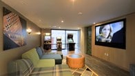 This single-floor entertainment lounge is thoughtfully designed to offer a relaxed yet engaging atmosphere. The space combines modern and retro elements seamlessly, creating an inviting environment for leisure and socializing. A standout feature is the collection of classic arcade game cabinets positioned near a floor-to-ceiling window, which floods the area with natural light and provides a view of the exterior surroundings. The large wall-mounted flat-screen television, framed by a distinctive stacked stone accent column, serves as a focal point for media viewing. Soft recessed lighting and wall sconces complement the natural light, creating a warm and well-lit ambiance throughout the day and evening. The seating area includes a plaid-upholstered sectional sofa and a unique cylindrical ottoman-style coffee table, offering comfortable and casual lounging options. The flooring transitions between wood-look planks and a patterned area rug, adding visual interest and defining the seating zone. The neutral gray walls and modern finishes contribute to a contemporary aesthetic, while the arcade machines introduce a fun, nostalgic character that makes this space both functional and distinctive.