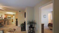 This single-floor residence presents a cohesive, comfortable layout with a neutral color palette of warm beige and cream tones carried consistently throughout the space. The main living area is anchored by a fireplace framed in dark stone or tile, serving as a natural focal point. Decorative elements, including potted ferns and sculptural accents, add organic warmth to the room.

The open sightlines from the living area extend toward what appears to be a kitchen or dining space, visible in the background and featuring light-toned cabinetry and what appears to be a refrigerator, suggesting a functional, well-connected layout. Carpeted flooring runs throughout the main living zones, contributing to a soft, inviting ambiance.

A hallway visible to the right leads to additional rooms, with a wooden bookcase or storage unit partially visible, indicating practical storage solutions within the home. Wall-mounted décor, including a circular mirror or art piece, adds dimension to the otherwise clean wall surfaces. The space benefits from ceiling-mounted ventilation and maintains a tidy, well-proportioned feel suited for everyday living.