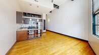 This loft-style unit in Winston-Salem's downtown core showcases industrial-chic character with soaring ceilings, exposed steel beams, and warm hardwood flooring throughout. The open-concept living and kitchen area is anchored by a large factory-style window wall that floods the space with natural light. The kitchen features dark wood cabinetry, granite countertops, stainless steel appliances, and pendant lighting over a freestanding island. The bedroom continues the warm hardwood aesthetic with clean white walls. The bathroom offers dark wood vanity cabinetry, granite countertops, a large mirror, and tile flooring, delivering a refined finish that complements the unit's urban character.