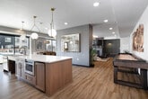 Located in Boise, Idaho, this thoughtfully designed community lounge offers a generous, open-plan layout that seamlessly blends a fully equipped kitchen with a comfortable living and gathering area. The kitchen is a standout centerpiece, featuring a long quartz-topped island with warm wood cabinetry, gold-accented bar seating, a marble-look tile backsplash, stainless steel appliances including a professional-grade range and French door refrigerator, and striking geometric globe pendant lights that add a modern, upscale touch. Large sliding glass doors along the perimeter flood the space with natural light and provide direct views of and access to the outdoor patio area. The living zone is anchored by a gas fireplace set against a sage-green paneled accent wall with a built-in bench, creating a cozy yet polished focal point. Light hardwood flooring runs throughout, unifying the space and enhancing its airy feel. Seating areas are thoughtfully arranged with a mix of leather lounge chairs, plush sofas, and a shuffleboard table, catering to both relaxed socializing and casual recreation. Decorative wall art and curated greenery add warmth and personality throughout. The overall ambiance is contemporary and inviting, balancing sophisticated design details with comfortable, functional gathering spaces.
