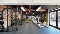 Located at 410 Court Street in Lynchburg, Virginia, this fitness center occupies a single open floor and offers a thoughtfully outfitted exercise space with a compelling blend of historic architectural character and modern gym amenities. Exposed brick walls and dark, heavy timber ceiling beams lend the space a distinctive industrial-rustic aesthetic, while rubber flooring throughout provides a durable, workout-ready surface. Bright overhead lighting ensures excellent visibility across the entire floor.

The cardio zone near the entrance features large glass doors and windows that invite natural light and open views of the surrounding streetscape, creating an airy atmosphere during workouts. Matrix-branded elliptical trainers and treadmills are arranged in a row, complemented by a stationary bike and other cardio machines. The strength training area is equally well-appointed, with a power rack loaded with weight plates, a full dumbbell rack, adjustable benches, a cable machine, and resistance equipment. A wall-length mirror in the strength zone enhances the sense of depth and allows for form monitoring. A secondary room at the rear provides additional space for specialized equipment. The combination of historic building character and comprehensive fitness equipment makes this an engaging and functional workout environment.