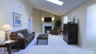 This single-floor space showcases an open and airy living area defined by its impressive vaulted ceilings that lend a sense of grandeur and spaciousness. A centrally positioned skylight floods the room with natural light, enhancing the warm, neutral tones of the walls and light-colored carpeted flooring throughout. The focal point of the living area is a classic white-mantled fireplace, framed by wall sconces that cast a soft, ambient glow and complemented by a wall-mounted flat-screen television above. The seating arrangement includes a plush sofa paired with a coordinating armchair, centered around a dark wood coffee table — offering a comfortable and cohesive gathering space. A substantial dark wood cabinet anchors one side of the room, providing functional storage while adding contrast to the lighter palette. Decorative touches such as potted greenery, floral accents, and tasteful accessories contribute to the polished, well-curated ambiance. The angled ceiling lines and thoughtful layout work together to create a space that feels both expansive and inviting, suitable for relaxed everyday living as well as entertaining.