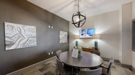 Located at 100 Myrtle St in Longwood, Florida, this single-floor space serves as a sophisticated meeting and lounge room designed for both collaborative gatherings and relaxed conversation. The room centers around a dark-toned round conference table surrounded by upholstered chairs, providing an intimate yet functional setting for small group meetings or discussions. A striking spherical metal chandelier hangs overhead, serving as a bold focal point and casting warm, ambient light throughout the space. The walls feature a rich charcoal-gray accent treatment complemented by neutral tones, and are adorned with abstract, earth-toned artwork that adds a contemporary aesthetic. A wall-mounted flat-screen television is positioned above a low wooden credenza, which is styled with teal ceramic table lamps, adding a pop of color to the overall neutral palette. Patterned carpet tiles in varying earth tones ground the space and contribute to its modern, professional character. A large interior window and glass-paneled sliding door connect the room to a brighter adjacent lounge area, allowing natural light to filter through and maintaining an open, connected feel within the broader facility. The overall ambiance is polished, welcoming, and well-appointed, making it an ideal space for meetings or community gatherings.