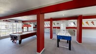 Located at 1900 Stevens Dr in Richland, Washington, this spirited two-floor entertainment space is designed with recreation and community in mind. The interior is defined by a bold red and gray color scheme, with wide horizontal crimson accent bands running throughout the walls, complemented by an extensive collection of collegiate pennants that lend the space a lively, team-spirited character.

The upper floor serves as the main living and entertainment level, offering generous open space with plush carpeting, recessed lighting, and large sliding glass doors that invite natural light. This floor features a full pool table with a branded overhead light fixture, a foosball table, a shuffleboard table, and chrome bar-height seating arrangements — creating a dynamic social hub ideal for gatherings and events.

A striking red-painted open staircase connects the two floors, leading down to the lower level, which continues the spirited aesthetic. This floor provides additional game room space, including an air hockey table and a ping pong table, with the same bold wall accents and pennant displays maintaining visual continuity throughout.

Both floors are finished with wall-to-wall carpeting, and the consistent design language across levels creates a cohesive, energetic atmosphere. The space is well-suited for community events, recreational use, or social entertainment.