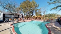 Located at 133 Lake Front Dr in Daphne, Alabama, this single-level outdoor amenity space centers around a generously sized freeform swimming pool with clear, blue-green water. The pool deck is finished with a classic brick-toned surround and is well-equipped for relaxation, featuring multiple lounge chairs and chaise seating arranged along the pool's perimeter. Vibrant orange market umbrellas are stationed throughout the deck, providing shaded seating areas ideal for warm Alabama days. The space is enclosed by a wrought iron fence, offering both security and an open, airy feel. Towering mature trees frame the area, creating a naturally canopied atmosphere that adds privacy and shade while enhancing the overall aesthetic. In the background, a multi-story residential building is visible, indicating the pool's role as a shared community amenity. The combination of well-maintained landscaping, ample seating, and the inviting pool makes this outdoor space a comfortable and functional retreat. Under clear blue skies, the setting evokes a relaxed, resort-like ambiance well-suited to the mild coastal climate of the Eastern Shore of Mobile Bay.