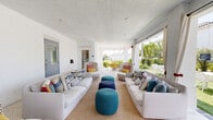 Located on Sumter Grove Circle in Naples, FL, this single-story community amenity space combines Florida-inspired architecture with a lively, resort-style aesthetic. The structure features crisp white exterior walls, elegant arched columns, and exposed dark wood beams overhead, complemented by a multi-toned paver terrace that extends across a generous covered outdoor area.

Multiple distinct lounge zones are thoughtfully arranged throughout the space. One area showcases a pair of oversized white sofas flanking colorful knit ottomans in teal and navy, all set atop a natural woven rug — ideal for casual gathering. A second seating vignette features rounded, sculptural chairs with vibrant red bases surrounding a cylindrical white coffee table, anchored by a bold teal accent wall adorned with a decorative mandala mural. Additional seating nooks with tall woven high-back chairs and teal pouf ottomans offer quieter spots for relaxation.

Open sightlines to the outdoors flood the space with natural light, framing views of swaying palm trees, a resort-style pool, and manicured green lawns beyond. Draping sheer curtains at open bays soften the transition between covered and open-air areas. Restroom facilities are conveniently accessible on-site. Throughout, the bright white palette is energized by pops of color, creating a welcoming and vibrant communal atmosphere.