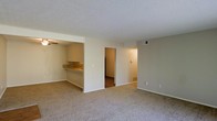 This single-floor residential unit offers a practical and functional layout designed to maximize open living space. The main living area is finished with wall-to-wall carpeting in a neutral tone, creating a comfortable and cohesive feel throughout. A textured popcorn ceiling adds a classic touch, while the walls are painted in a soft, warm beige that lends a welcoming ambiance to the space.

A defining feature of the layout is the pass-through opening that connects the living area to the kitchen, allowing natural sightlines and an open feel between the two spaces. The kitchen features a countertop bar ledge, ideal for casual dining or food prep, and is equipped with a ceiling fan for added ventilation. The countertops display a warm, earthy tone that complements the overall color palette.

Ventilation is well-addressed throughout, with visible HVAC vents integrated into both the walls and floors, ensuring climate control across the unit. A doorway from the main living area leads to an adjacent room, suggesting a straightforward and accessible floor plan. The space is clean, uncluttered, and move-in ready, making it a versatile canvas suitable for a variety of furnishing styles and personal preferences.