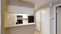 This single-story space centers around a practical kitchen designed for efficient use. The kitchen features white upper and lower cabinetry that extends to the ceiling, maximizing storage capacity. A peninsula counter with a light-toned surface extends outward, creating a natural divider between the kitchen and the adjacent area while offering additional workspace or casual seating potential. Black appliances, including an electric range with an overhead exhaust hood and a full-size refrigerator, provide a notable contrast against the white cabinetry. Light-toned countertops complement the overall neutral color palette. The flooring consists of light wood-style planks that run through the kitchen and into the adjoining hallway or room beyond. The walls and ceilings feature a textured finish consistent throughout the visible areas, contributing to the space's uniform aesthetic. A hallway adjacent to the kitchen leads to additional areas of the unit, with two white doors visible, suggesting access to storage or separate rooms. The overall color scheme relies on neutral beige and white tones, creating a clean and straightforward interior environment.