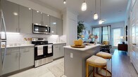 This well-designed single-floor residence combines modern aesthetics with functional living spaces. The kitchen is a standout feature, showcasing sleek grey cabinetry with a high-gloss finish, stainless steel appliances including a range, microwave, refrigerator, and dishwasher, and a marble-look tile backsplash. A peninsula island with a white quartz countertop offers additional workspace and seating with stylish bar stools, while pendant lighting adds a refined touch overhead. Light wood-style flooring flows throughout, enhancing the open and airy feel.

The living area is bright and inviting, with large windows dressed in tailored blinds and drapes that allow generous natural light. The space carries a contemporary character accented by bold artwork and decorative wall pieces that lend personality to the room.

The bedroom is generously sized, featuring large windows that bathe the room in natural light, complemented by crown molding and light wood-style flooring. A second bedroom provides equally comfortable accommodations with similar finishes and ample natural light.

The bathroom is thoughtfully laid out with grey cabinetry, a white countertop, a wide vanity mirror, and wood-look tile flooring. A spacious walk-in closet with built-in shelving and hanging rods is conveniently accessible directly from the bathroom, offering excellent storage throughout this well-appointed residence.