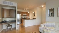 This single-floor space is designed with an open-concept layout that seamlessly connects the kitchen and living areas, creating a cohesive and functional environment. The kitchen is a standout feature, outfitted with stainless steel appliances, including a refrigerator, stove, and built-in microwave. Warm-toned wooden cabinetry provides ample storage, complemented by dark countertops that add contrast and visual interest. A white peninsula island extends into the space, offering additional prep space and serving as a natural divider between the kitchen and living area. Pendant lighting hangs above the island and a track lighting system illuminates the kitchen, ensuring well-lit work surfaces. The living area flows naturally from the kitchen, anchored by light hardwood flooring that extends throughout, adding warmth and continuity. Neutral cream-toned walls keep the space feeling bright and airy. A doorway visible from the main area hints at additional rooms beyond, suggesting further functional space. Overall, the layout balances practicality and comfort, making it a well-appointed and inviting single-floor residence.