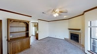 This two-floor home offers a functional and comfortable layout designed for everyday living. The main living area is a welcoming space finished with plush carpeting and neutral-toned walls accented by warm wood crown molding that frames the room with a classic touch. A ceiling fan with integrated lighting anchors the room from above, providing both airflow and illumination. A standout feature of this living space is the tile-surround fireplace, complete with a raised hearth and a wooden mantel, serving as a natural focal point. Built-in wooden shelving and cabinetry along one wall offer generous display and storage space while adding a traditional, homey character to the room. An open passageway connects the living area to the kitchen, which features wooden cabinetry and includes a full-size refrigerator, allowing for a convenient flow between spaces. Recessed lighting in the transitional area further enhances the connectivity between rooms. The combination of warm wood tones, soft carpeting, and classic architectural details throughout creates an inviting and lived-in atmosphere across both floors of this home.