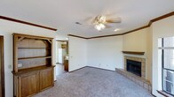 This two-floor home offers a functional and comfortable layout designed for everyday living. The main living area is a welcoming space finished with plush carpeting and neutral-toned walls accented by warm wood crown molding that frames the room with a classic touch. A ceiling fan with integrated lighting anchors the room from above, providing both airflow and illumination. A standout feature of this living space is the tile-surround fireplace, complete with a raised hearth and a wooden mantel, serving as a natural focal point. Built-in wooden shelving and cabinetry along one wall offer generous display and storage space while adding a traditional, homey character to the room. An open passageway connects the living area to the kitchen, which features wooden cabinetry and includes a full-size refrigerator, allowing for a convenient flow between spaces. Recessed lighting in the transitional area further enhances the connectivity between rooms. The combination of warm wood tones, soft carpeting, and classic architectural details throughout creates an inviting and lived-in atmosphere across both floors of this home.