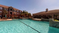This outdoor pool area offers a resort-style experience centered around a generously sized swimming pool with crystal-clear turquoise water. The pool is framed by tiled water feature walls that cascade water from elevated ledges, adding both visual interest and a soothing ambiance. Surrounding the pool, the stone-tiled deck provides ample space for lounging, and a robust wooden pergola offers shaded seating with outdoor furniture for relaxation.

A covered patio runs along one side of the space, supported by brick columns and equipped with ceiling fans to maintain airflow during warm weather. Stone tile flooring extends throughout this covered area, which features outdoor dining tables and chairs, making it suitable for casual gatherings. Access to the pool area is controlled through a secured metal gate entry.

The architectural design of the surrounding buildings is traditional in style, constructed with warm brick exteriors and pitched rooflines. Mature palm trees and manicured landscaping are integrated throughout, enhancing the tropical atmosphere. The entire space is open to the sky, allowing abundant natural sunlight to illuminate the area, while the surrounding multi-story brick buildings provide a sense of enclosure and privacy, creating an inviting and well-appointed outdoor retreat.
